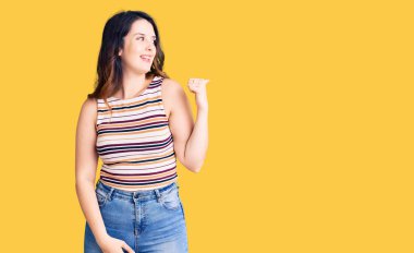 Güzel, genç, esmer, gündelik kıyafetler giyen mutlu yüzüyle gülümseyen ve başparmağıyla yan tarafı gösteren bir kadın.. 