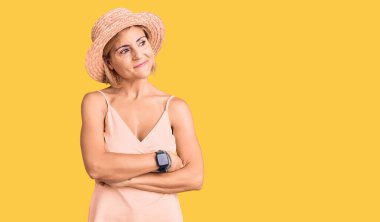 Sarışın, yaz şapkası takan, kolları çapraz ve kendinden emin genç bir kadın. 