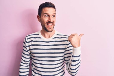 Sıradan çizgili süveter giyen genç yakışıklı, pembe arka planda dikiliyor, baş parmağını yana kaldırıyor ve ağzı açık gülümsüyor.