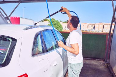 Orta yaşlı yakışıklı, günlük kıyafetler giyen ve mutlu gülümseyen güneş gözlüklü bir adam. Yüzünde gülümsemeyle ayakta durup su tabancası kullanarak araba yıkamak..
