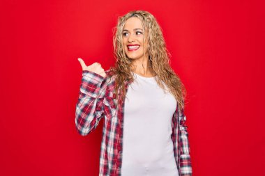 Genç, sarışın, sıradan bir gömlek giyen, kırmızı arka planda duran mutlu yüzüyle gülümseyen ve baş parmağıyla yan tarafı gösteren bir kadın..