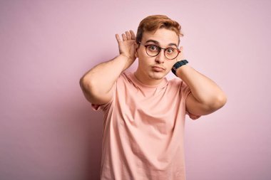 Genç, kızıl saçlı, günlük tişört giyen, pembe arka planda duran, avuçlarında alaycı ve kuşkucu ellerle tavşan kulağı hareketi yapan genç bir adam. Paskalya tavşanı kavramı.