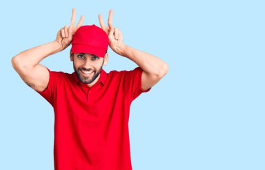 Sakallı, teslimat üniforması giyen, komik ve deli pozu veren, parmakları tavşan kulaklı, neşeli gülümseyen genç yakışıklı bir adam. 