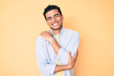 Sarı arka planda duran yakışıklı İspanyol adam kendine sarılıyor mutlu ve pozitif, kendinden emin gülümsüyor. Sevgi ve özgüven. 