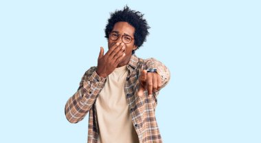 Afro saçlı, sıradan giyinmiş ve gözlüklü yakışıklı Afro-Amerikalı adam sana gülüyor, eli ağzında kamerayı işaret ediyor, utanç ifadesi takınıyor. 