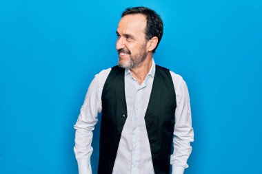 Zarif gömlek ve yelek giyen orta yaşlı yakışıklı bir adam mavi arka planda, yan tarafa bakıyor, rahat profil pozu veriyor doğal bir yüzü ve kendine güvenen bir gülümsemesi var..