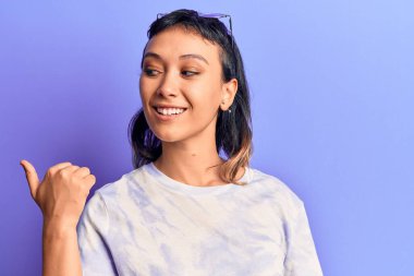Gündelik kıyafetler giyen, gülümseyen, gülen yüzlü genç bir kadın başparmağıyla yan tarafa bakıyor ve işaret ediyor.. 