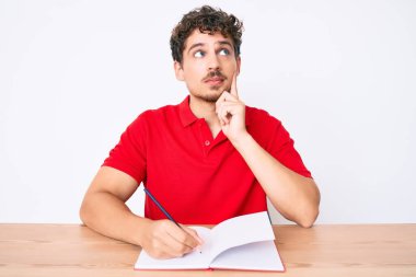 Kafkasyalı, kıvırcık saçlı genç adam masaya oturmuş defter yazıyor. Ciddi bir yüz ifadesi var. Çene üstüne çeneyle soru sormayı düşünüyor. 