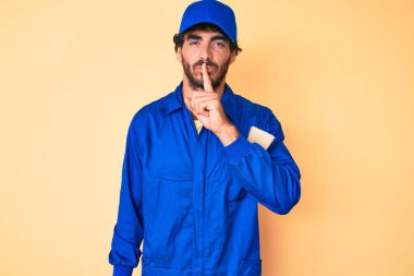 Kıvırcık saçlı, ayı kılıklı, işçi tulumu giyen yakışıklı genç adam, parmakları dudaklarında sessiz olmak istiyor. sessizlik ve gizli konsept. 