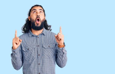 Gündelik giysiler giyen genç Arap şaşırmış ve şaşırmış bir şekilde yukarı bakıyor ve parmaklarıyla işaret ediyor ve kollarını kaldırıyordu.. 