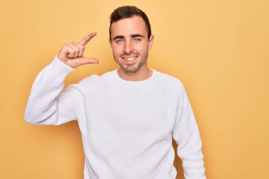Mavi gözlü, sıradan bir süveter giyen, sarı arka planda duran, gülümseyen, kendine güvenen, el hareketi yapan, küçük boy işareti yapan, parmaklarıyla kameraya bakan genç, yakışıklı bir adam. Ölçü kavramı.