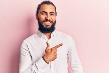 Yüzünde mutlu ve doğal bir ifade olan, elleri ve parmaklarıyla işaret eden, güler yüzlü, gündelik giysiler giyen genç bir Arap. 