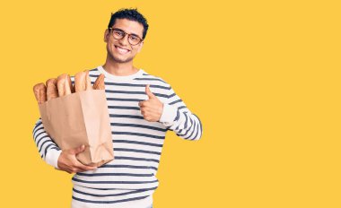 İspanyol yakışıklı genç adam elinde kağıt torbayla mutlu ve pozitif gülümsüyor, başparmağıyla mükemmel ve onay işareti yapıyor. 