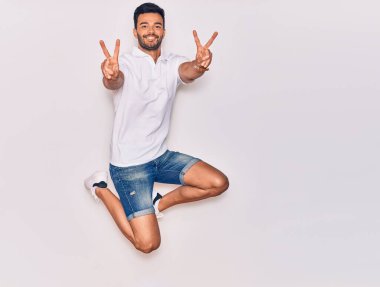 Genç, yakışıklı, sıradan kıyafetler giyen, mutlu gülümseyen bir İspanyol. Yüzünde gülümsemeyle zıplıyor zafer işareti yapıyor parmakları izole beyaz arka planda.