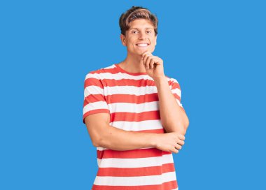 Gündelik giysiler giyen, kameraya kendinden emin, kolları çapraz ve çenesi havada gülümseyen genç yakışıklı bir adam. Olumlu düşünmek. 