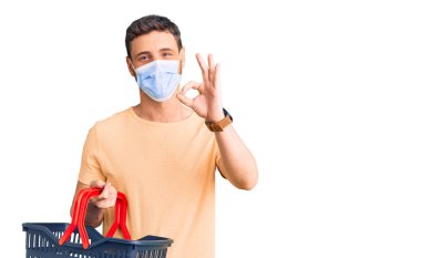 Alışveriş sepeti ve sağlık maskesi takan yakışıklı genç adam parmaklarıyla imza atıyor, gülümsüyor ve mükemmel bir sembol oluşturuyor. 