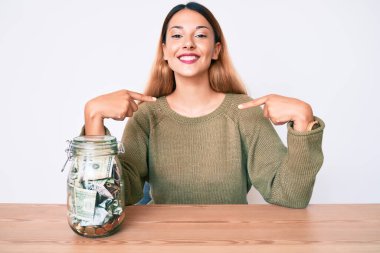 Masada bir kavanoz dolusu parayla oturan genç esmer kadın. Kendine güvenen, yüzünde gülümseme olan, kendini gururlu ve mutlu gösteren bir kadın.. 