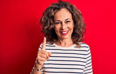 Orta yaşlı, esmer, çizgili tişört giyen güzel bir kadın kırmızı arka planda duruyor. Bir fikir veya soruyla gülümsüyor ve mutlu bir yüzle işaret ediyor.