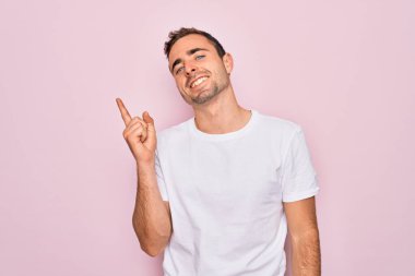 Yakışıklı, mavi gözlü, sıradan beyaz tişörtlü, pembe arka planda duran, yüzünde kocaman bir gülümseme olan, eli ve parmağı kameraya bakan bir adam..