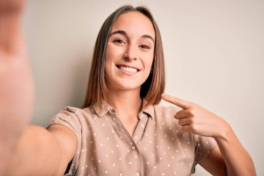 Yazlık tişört giyen genç ve güzel bir kadın beyaz arka planda kamerayla selfie yapıyor. Elle ve parmakla işaret ederek çok mutlu.