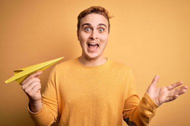 Genç ve yakışıklı kızıl saçlı adam izole edilmiş sarı arka plan üzerinde kağıt uçak tutuyor mutlu ve heyecanlı, kazanan yüz ifadesi zaferi büyük bir gülümsemeyle kutluyor ve ellerini kaldırıyor.