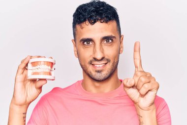 Elinde takma diş tutan genç İspanyol bir adam bir fikir veya soruyla gülümsüyor ve mutlu bir yüzle işaret ediyor. 