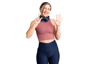 Güzel beyaz kadın spor kıyafetleri giyiyor ve kulaklık takıyor. Yedi numaralı parmaklarıyla işaret ediyor. Kendinden emin ve mutlu bir şekilde gülümsüyor.. 