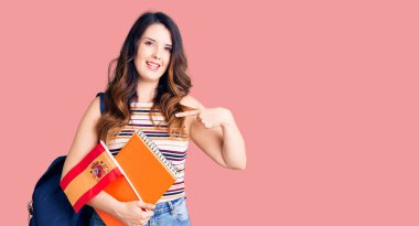 Güzel, genç, esmer, değişim öğrencisi, elinde İspanya bayrağı, gülümseyen mutlu ve gururlu birini işaret ediyor. 