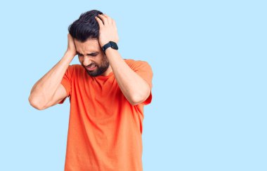 Sakallı, gündelik tişörtlü, baş ağrısı çeken, çaresiz ve stresli genç bir adam. Ağrı ve migren yüzünden. Eller başının üstünde.. 