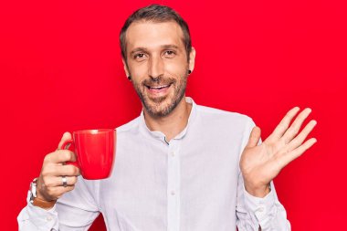 Genç yakışıklı adam kahveyle başarıyı kutluyor mutlu bir gülümsemeyle ve kazanan yüz ifadesiyle elini kaldırarak. 