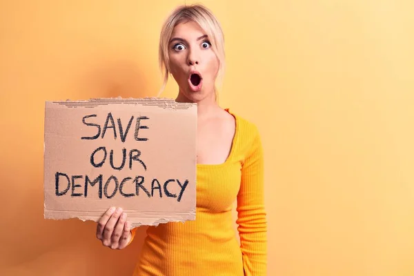 Young blonde woman asking for politics holding banner with save our democracy message scared and amazed with open mouth for surprise, disbelief face