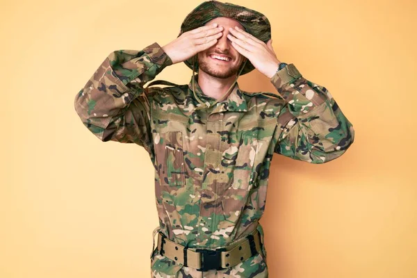 Young caucasian man wearing camouflage army uniform covering eyes with hands smiling cheerful and funny. blind concept.