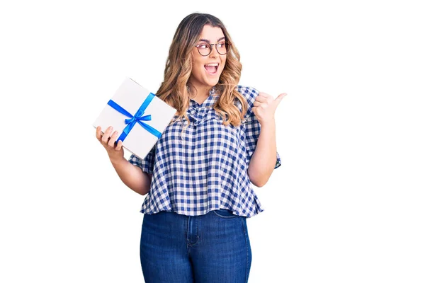 Joven Mujer Caucásica Sosteniendo Regalo Señalando Pulgar Hacia Lado Sonriendo — Foto de Stock