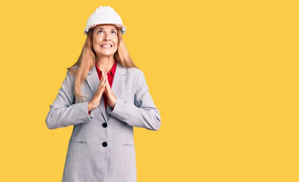 Beautiful young woman wearing architect hardhat begging and praying with hands together with hope expression on face very emotional and worried. begging.