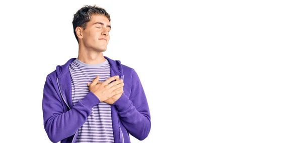 年轻英俊的男子穿着休闲的紫色运动衫 双手放在胸前笑着 闭上了眼睛 脸上挂着感激的手势 健康概念 — 图库照片