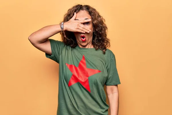 Middle age beautiful woman wearing t-shirt with red star revolutionary symbol of communism peeking in shock covering face and eyes with hand, looking through fingers afraid