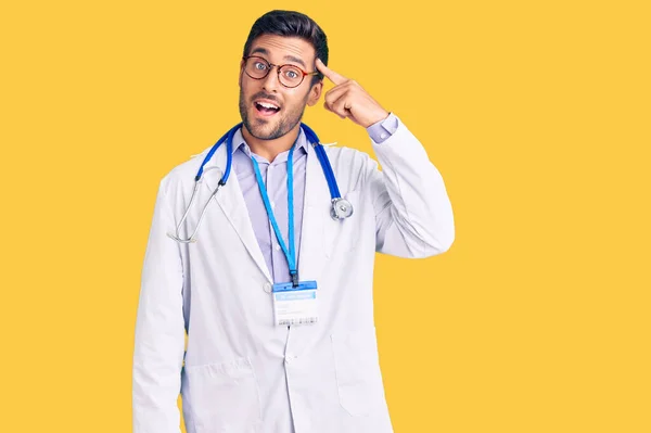 医者の制服を着た若いヒスパニック系の男と一本の指で頭を指して笑顔聴診器 素晴らしいアイデアや考え 良い記憶 — ストック写真