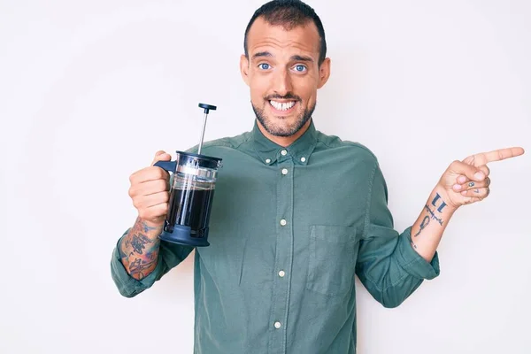 Young handsome man with tattoo holding french coffee maker smiling happy pointing with hand and finger to the side