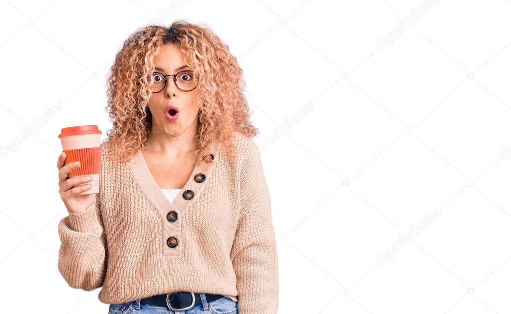 Young blonde woman with curly hair wearing glasses and drinking a cup of coffee scared and amazed with open mouth for surprise, disbelief face 