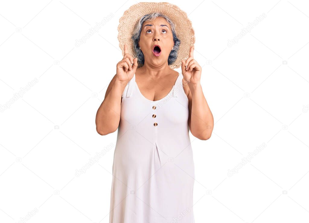 Senior woman with gray hair wearing summer hat and summer clothes amazed and surprised looking up and pointing with fingers and raised arms. 