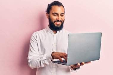 Genç Arap adam dizüstü bilgisayarı tutuyor. Pozitif ve mutlu görünüyor. Kendine güvenen bir gülümsemeyle ayakta duruyor ve dişlerini gösteriyor. 