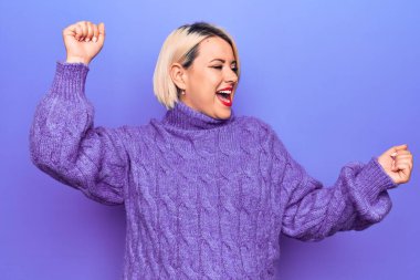 Güzel sarışın ve cüsseli bir kadın mor arka planda balıkçı yaka kazak giyiyor mutlu ve neşeli dans ediyor, hareketli ve kendinden emin bir şekilde müzik dinliyor.