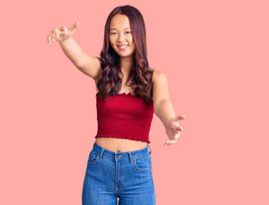 Gündelik giysiler giymiş genç, güzel Çinli bir kız kameraya bakıyor ve sarılmak için kollarını açmış gülümsüyor. Mutluluğu kucaklayan neşeli bir ifade.. 