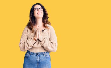 Güzel, genç, esmer bir kadın günlük kıyafetler ve gözlük takıyor. Elleriyle yalvarıyor ve dua ediyor. Yüzünde umut ifadesi var. Yalvarıyor. 