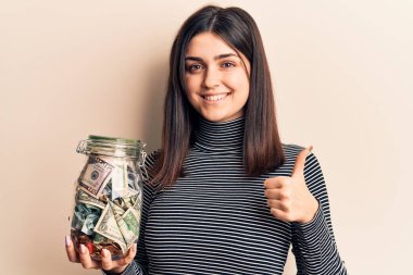 Elinde bahşiş kavanozu tutan genç ve güzel bir kız mutlu ve pozitif gülümsüyor, başparmağıyla mükemmel ve onay işareti yapıyor. 