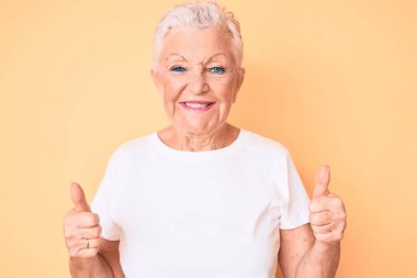 Son sınıf güzeli, mavi gözlü, gri saçlı, sarı arka plan başarı işareti üzerine klasik beyaz tişört giyen, el hareketleriyle olumlu hareket eden, mutlu ve gülümseyen bir kadın. Neşeli ifade ve kazanan jest. 