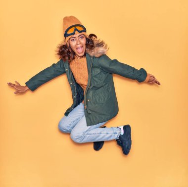 Kışlık giysiler giyen genç, güzel Latin kadın mutlu gülümsüyor. Yüzünde ve kollarında tebessümle, izole edilmiş sarı arka planda atlıyor.