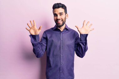 Sakallı, sıradan tişörtlü, on numaralı parmaklarıyla onu gösteren, kendinden emin ve mutlu gülümseyen genç bir adam.. 