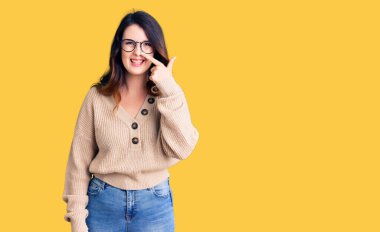 Güzel, genç, esmer bir kadın, günlük kıyafetler ve gözlük takıyor, parmakları burnunda, neşeli bir şekilde gülümsüyor. güzellik konsepti 