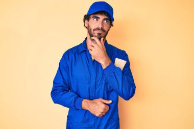 Kıvırcık saçlı, ayı kılıklı, eli çenesinde inşaatçı tulumu giyen, soru soran, düşünceli, yakışıklı bir genç. Düşünceli bir yüzle gülümsüyor. şüphe kavramı. 